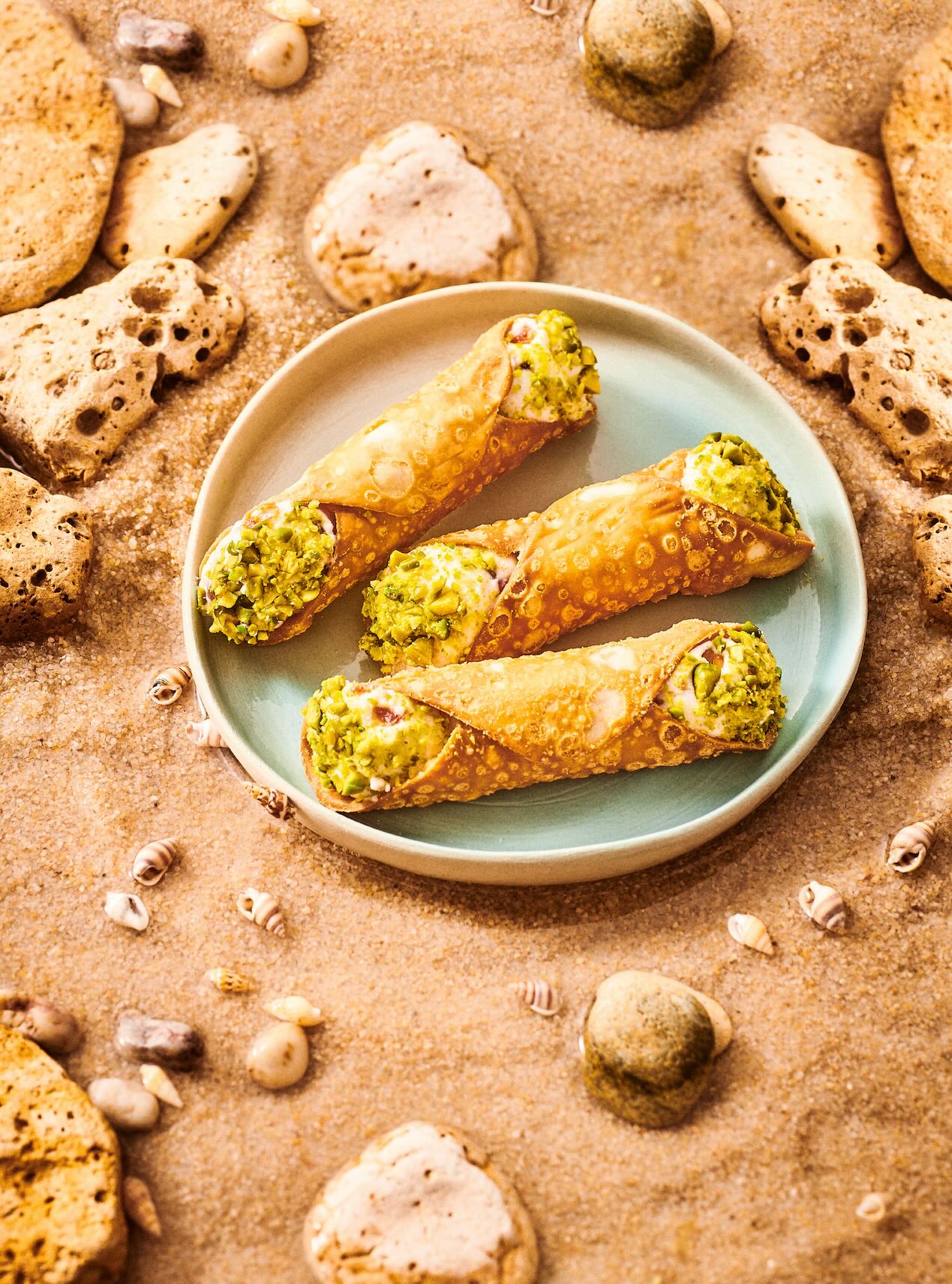 Three ricotta and pistachio cannoli on a blue plate, surrounded by sand, rocks, and small seashells
