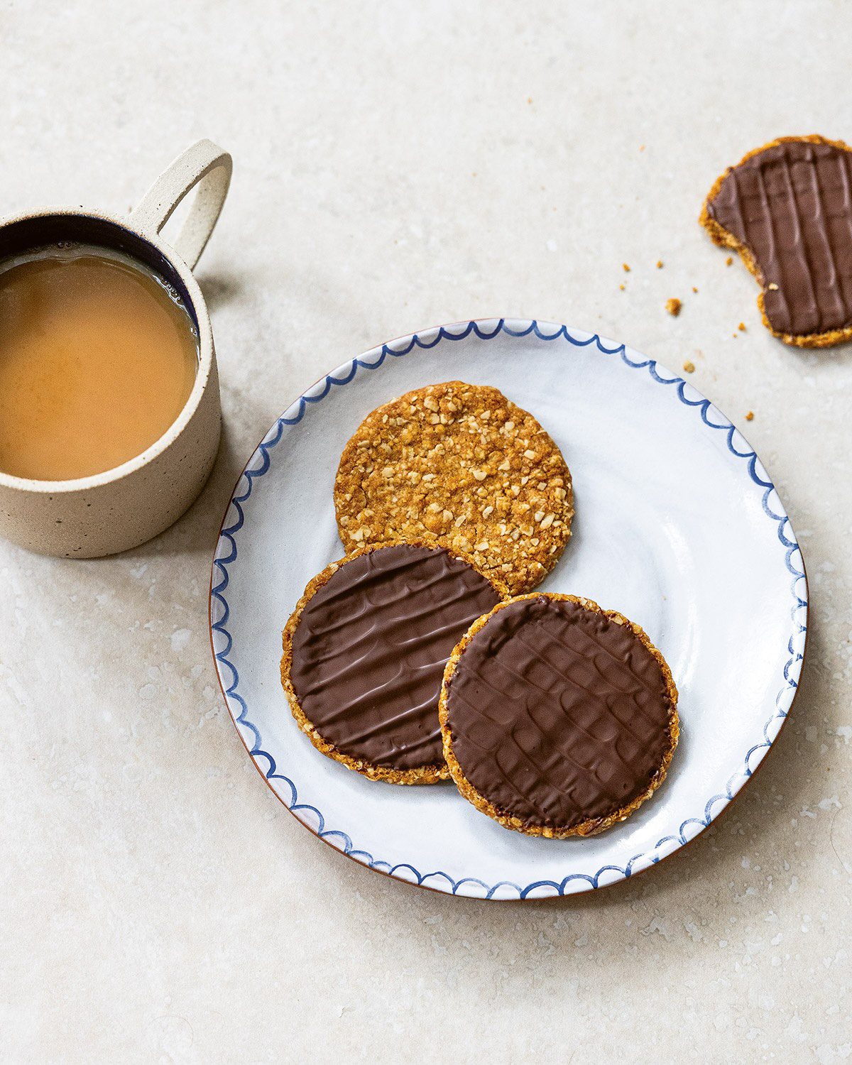 Three knobbly chocolate oat cookies on a ceramic plate beside a mug of tea, with one bitten cookie