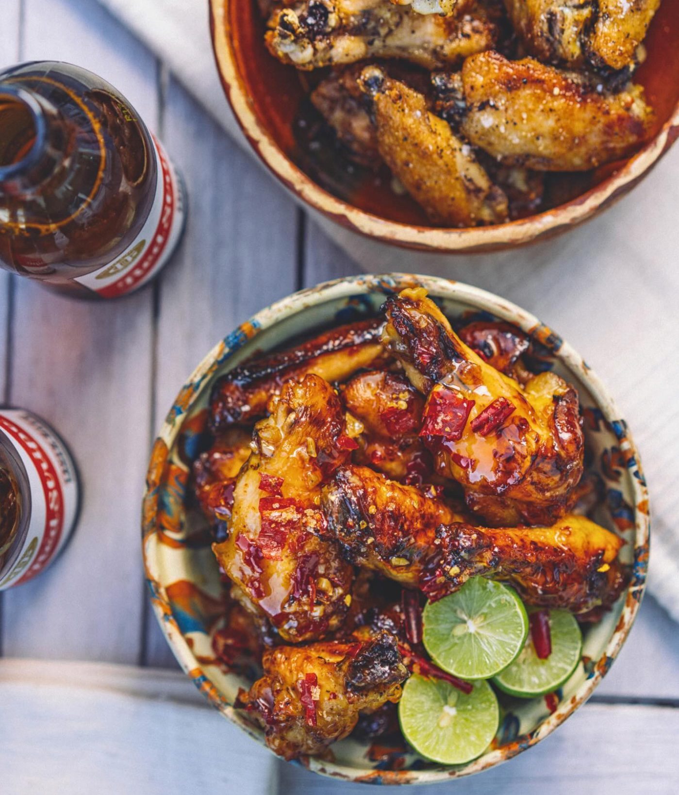 Two bowls of chicken wings, one spicy with lime slices, and two beer bottles on a wooden table.