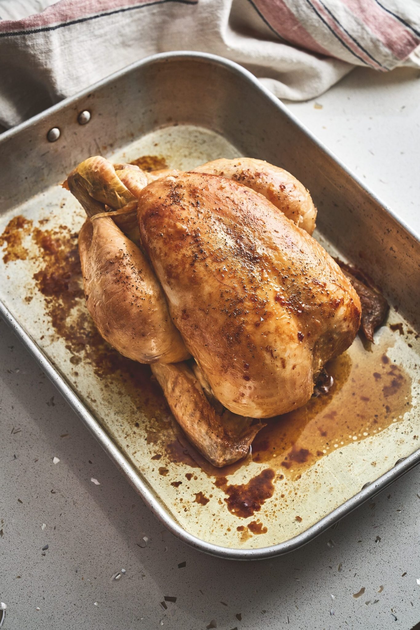 Golden-brown roast chicken resting in a metal roasting pan, surrounded by flavorful juices.