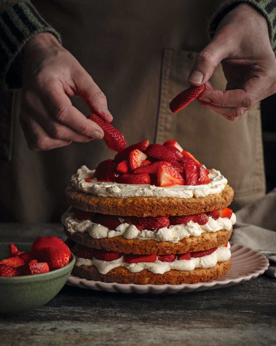 Hands placing fresh strawberries on top of a layered strawberry shortcake filled with whipped cream.