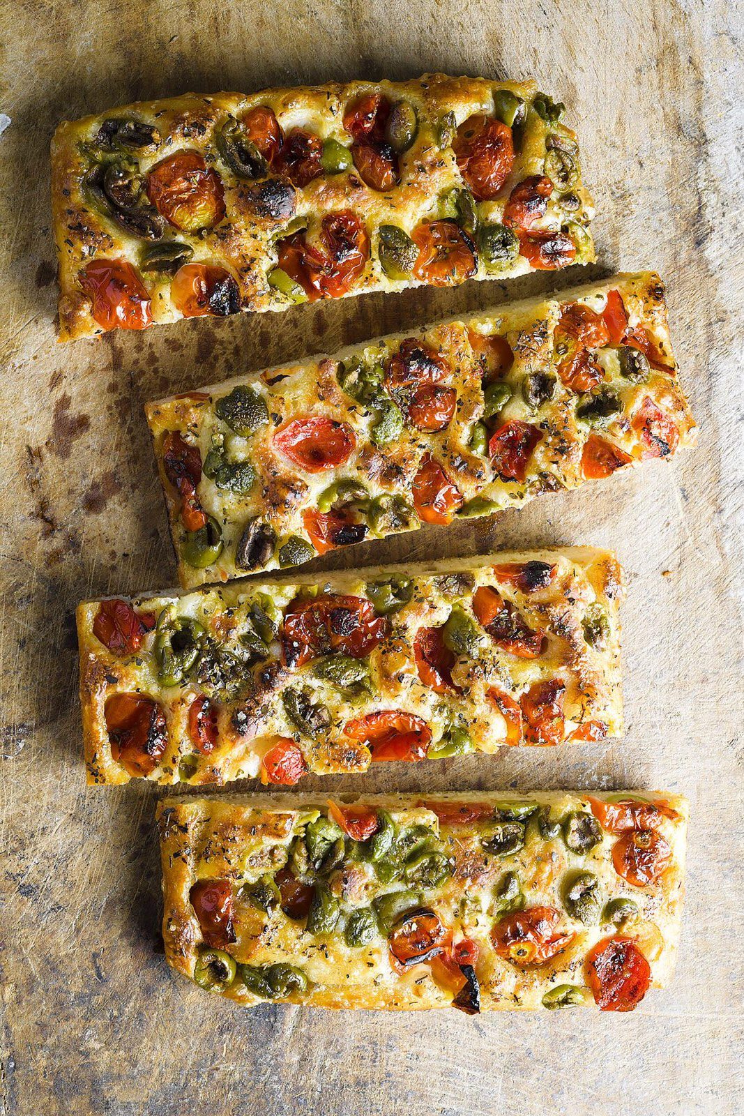 Slices of golden focaccia topped with tomatoes, olives, and herbs on a wooden surface.