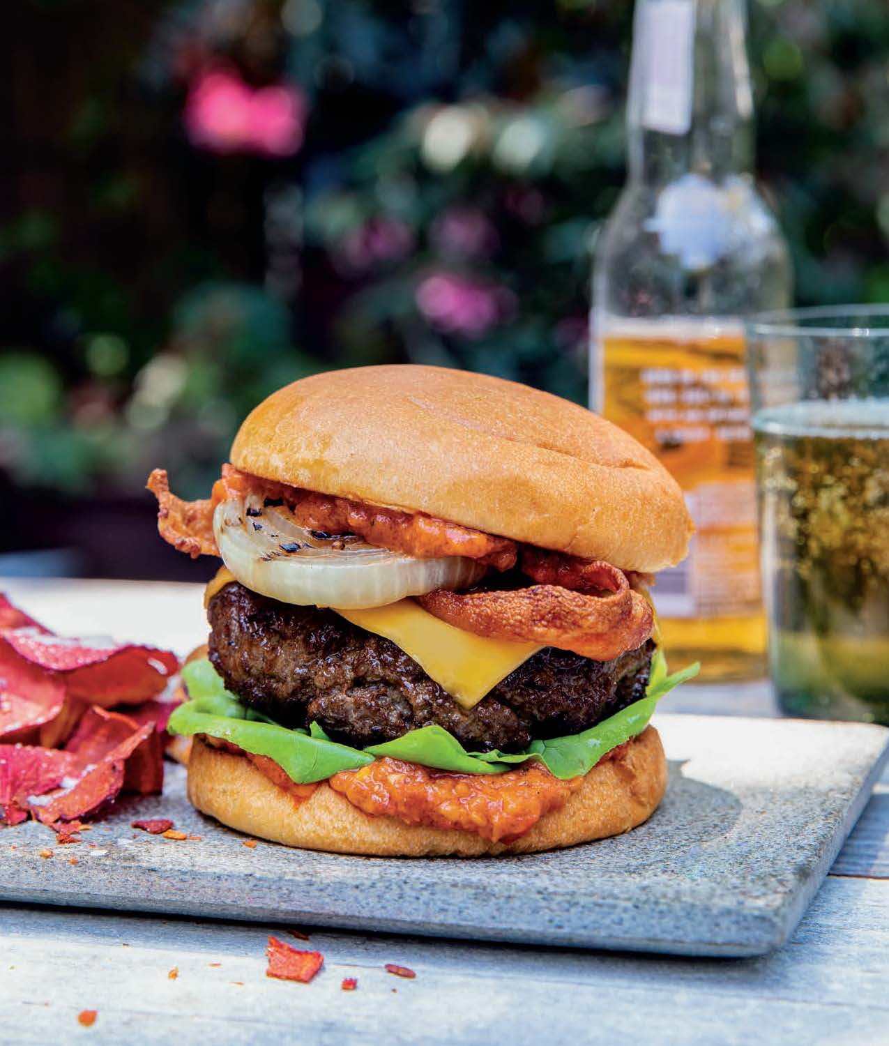 A gourmet burger with cheese, served with reddish chips and beer outdoors.