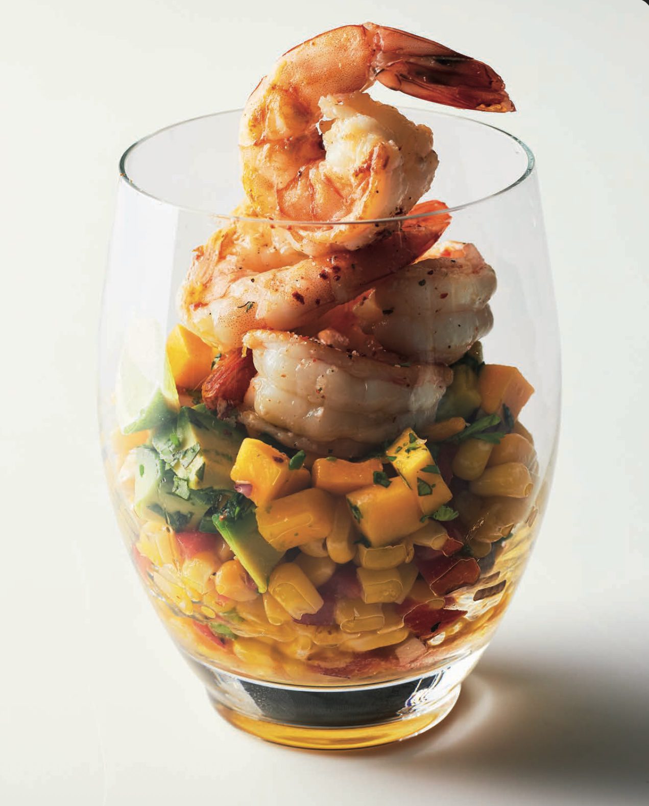 Grilled shrimp stacked over a colourful mango, avocado, and corn salad in a clear glass.